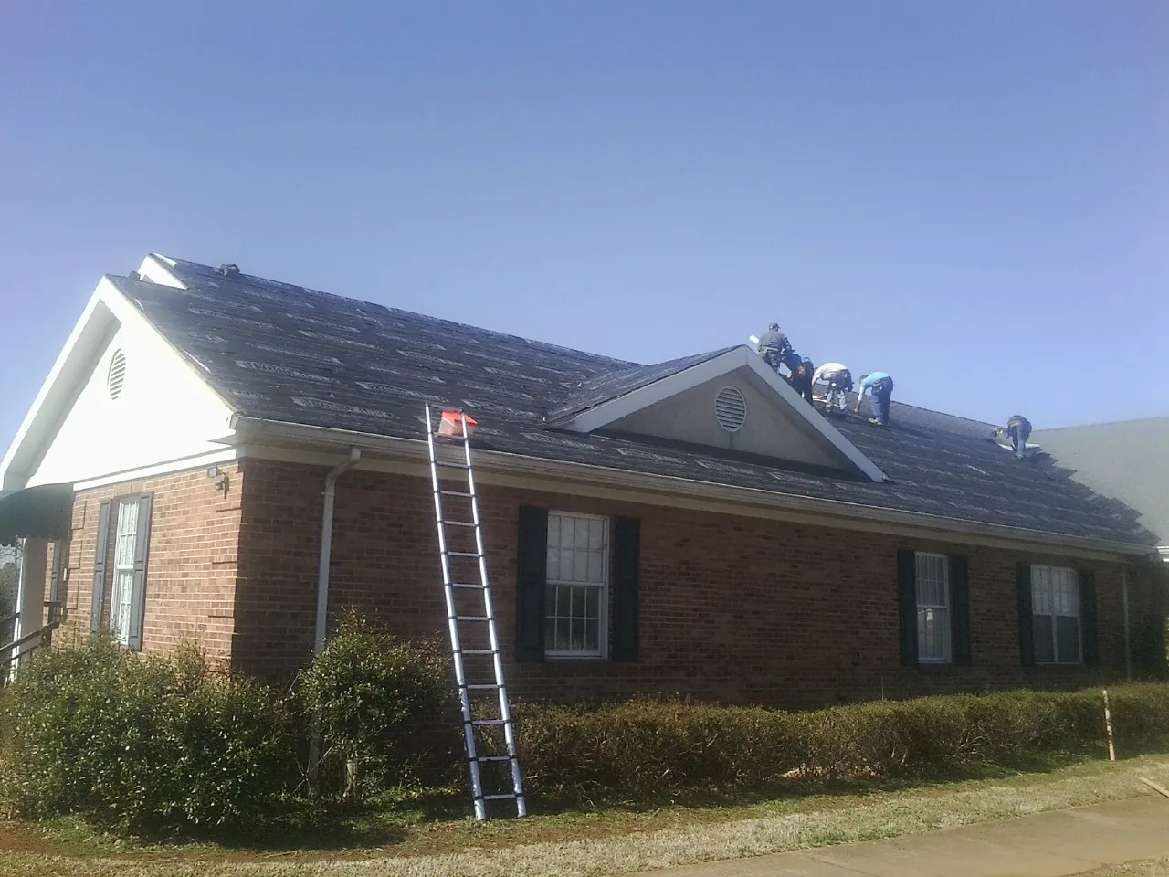 Crew tearing shingles off a brick ranch roof with a ladder set — Emergency Roof Repair work by our crew in Oldenburg