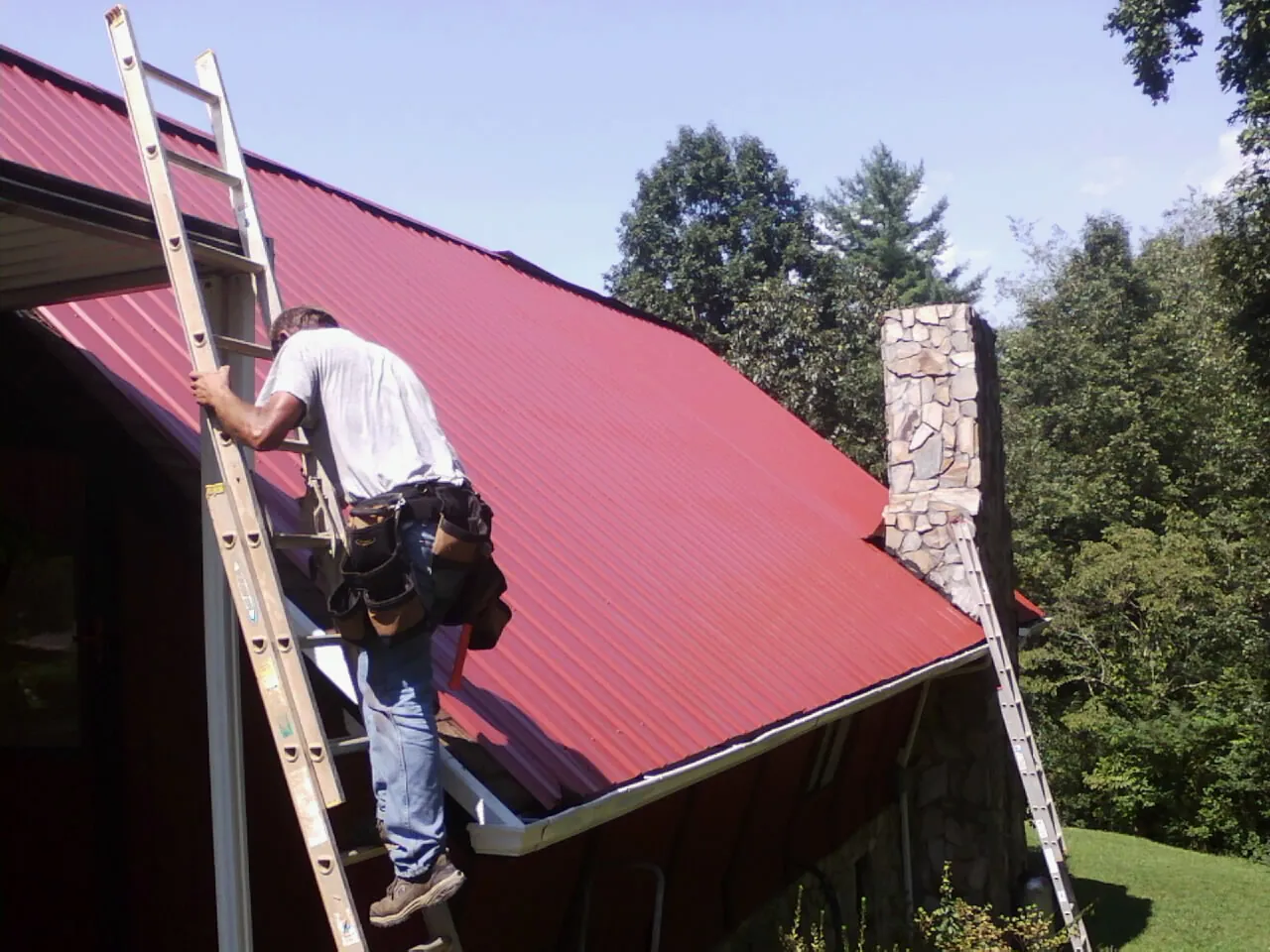 Roofer on a ladder beside a red metal roof and stone chimney — Soffit & Fascia Repair by Roofing Brothers in Oldenburg